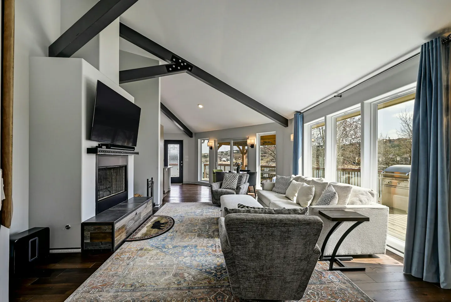 Bright living room with dark wood floors, exposed beams, and a stone fireplace with a TV above. Large windows with blue curtains.