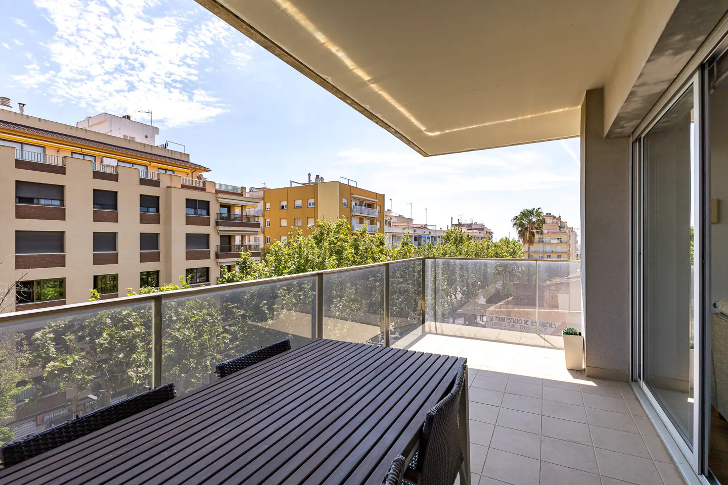 A balcony with a dark wood table and chairs overlooks a city with trees under a blue sky.