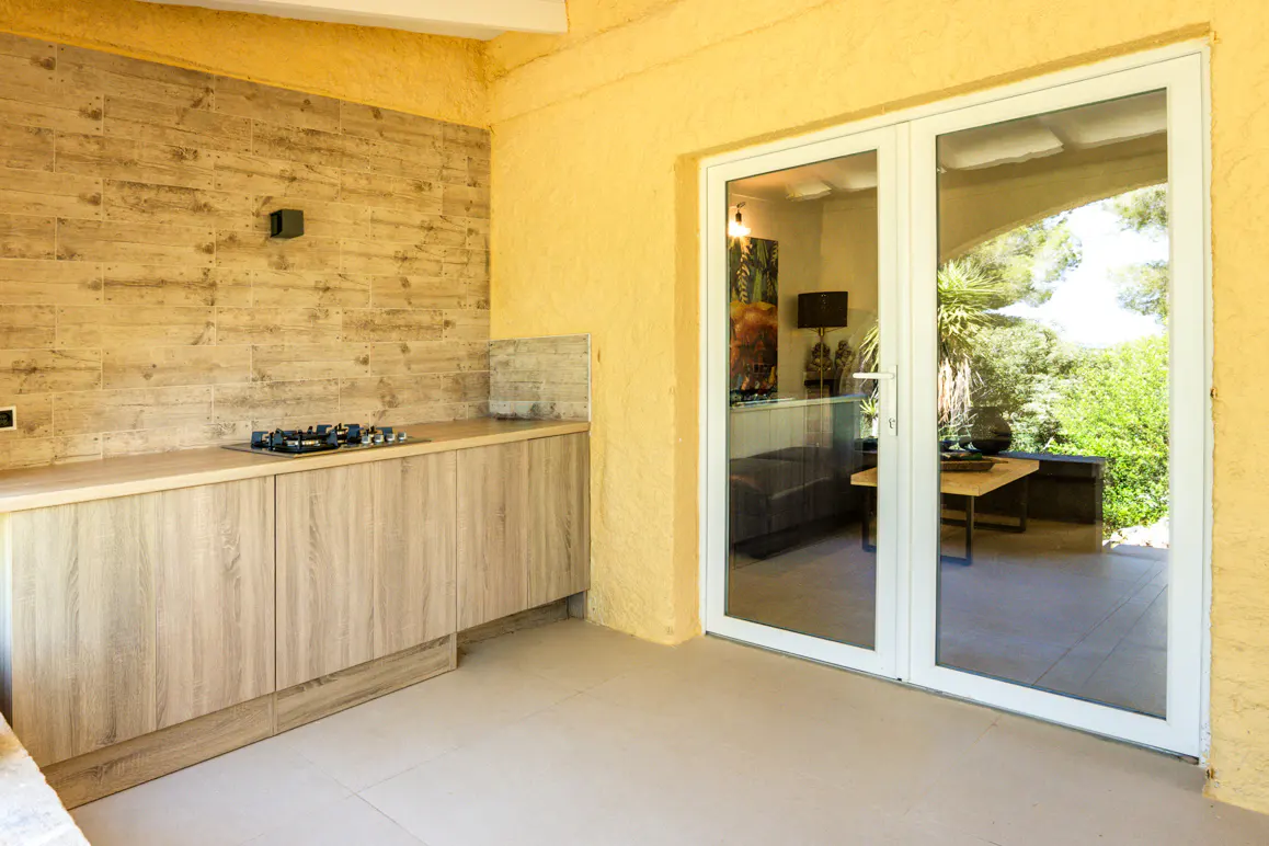 Outdoor kitchen with wood cabinets, gas cooktop, and textured yellow walls. White framed glass doors lead to a patio and garden.
