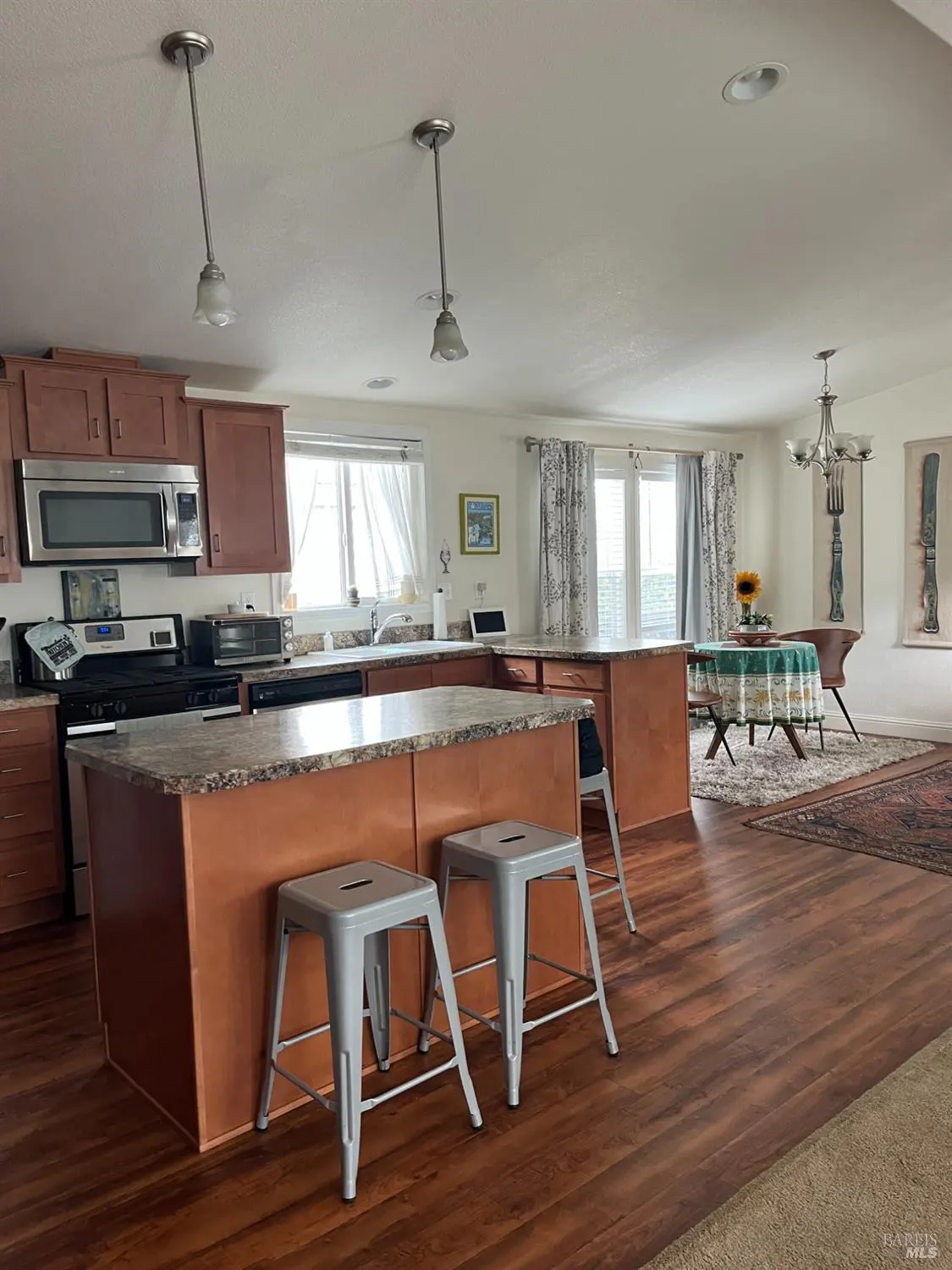 A bright, open kitchen with wood floors, brown cabinets, and two gray stools at the island. A dining area is visible in the background.