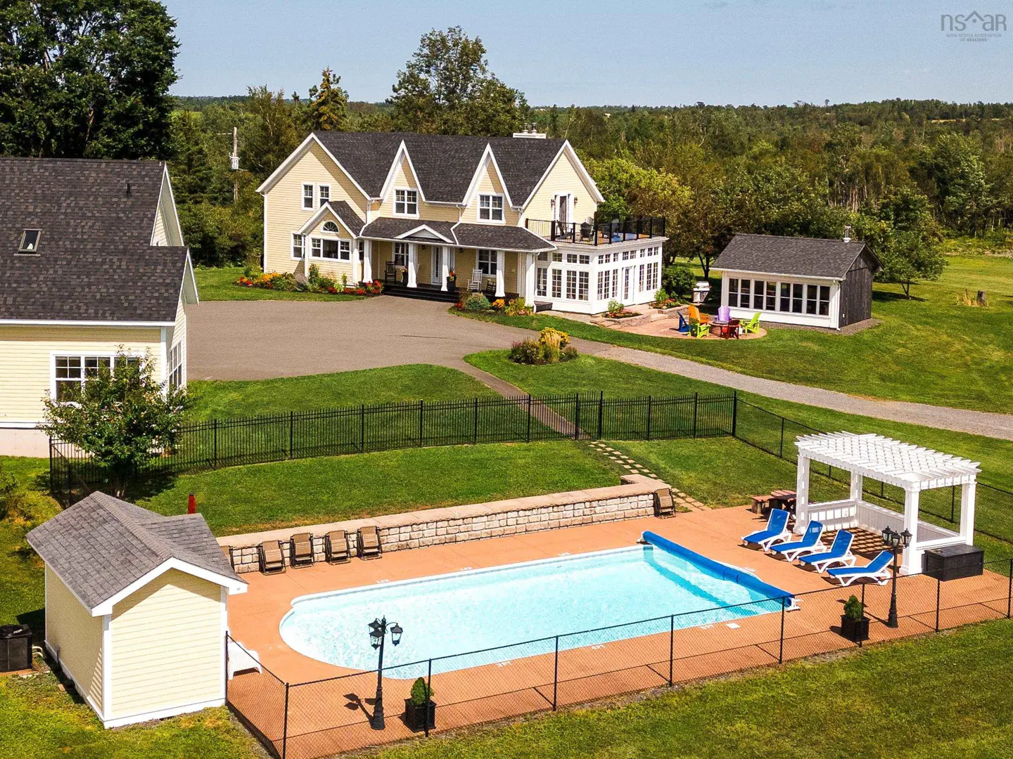 Aerial view of a yellow two-story house with a pool, pergola, and green lawn. Trees and a blue sky are in the background.