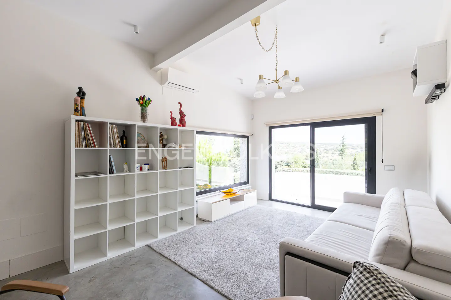 Bright living room with white walls, a large window, a white sofa, and a white bookshelf filled with books and decorative items.