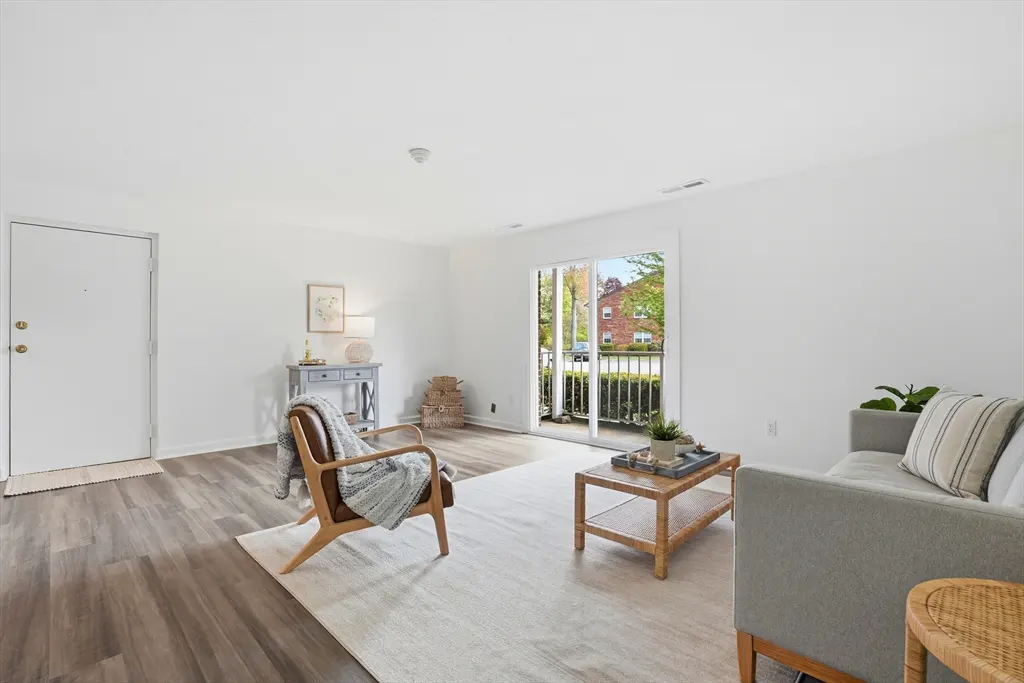 Bright living room with white walls, wood floors, and a sliding glass door to a balcony. A gray sofa and wood chair sit on a light rug.