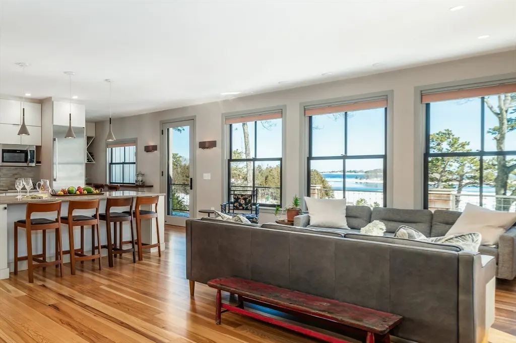 Open-concept living space with wood floors, a gray sofa, and a kitchen island with bar stools. Large windows offer a view of a lake.