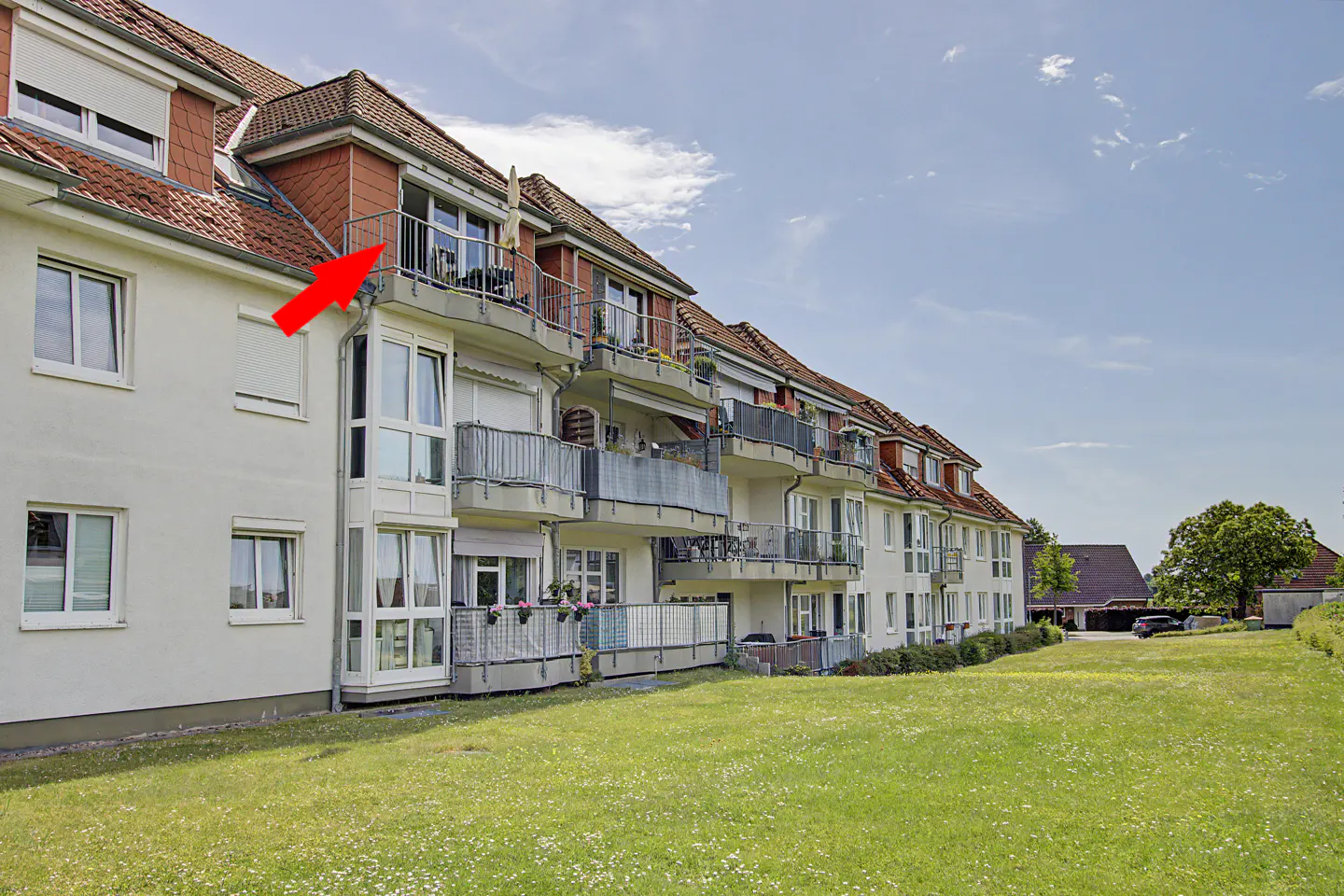 A red arrow points to a top-floor balcony on a multi-story apartment building with a grassy lawn under a blue sky.
