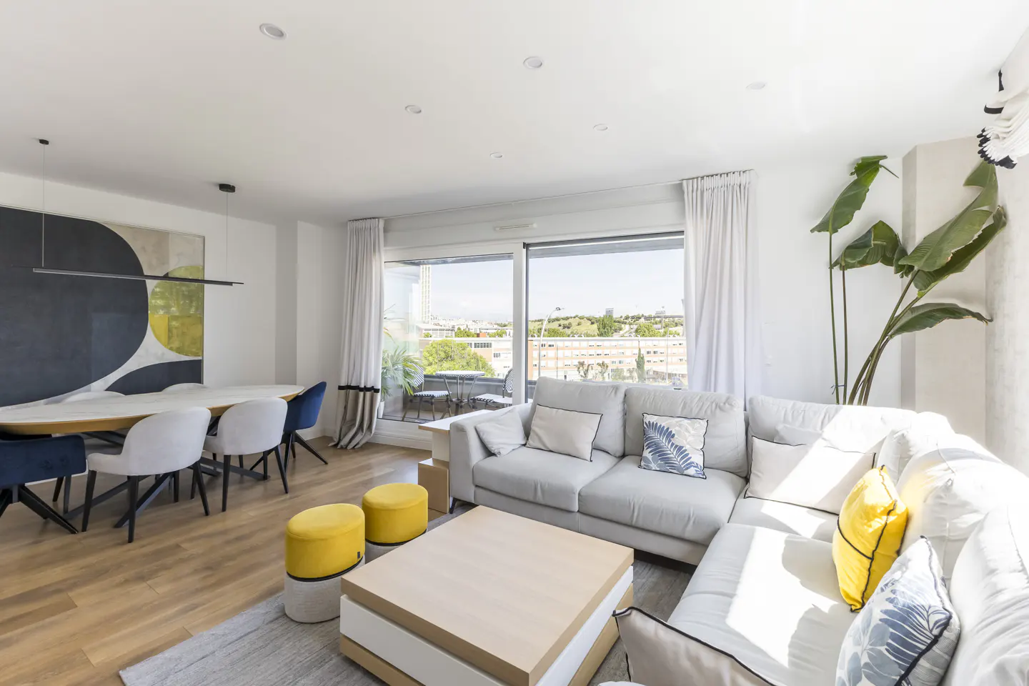 Bright, modern living room with a gray sectional sofa, wood floors, and a dining area with a large abstract painting. Balcony view.