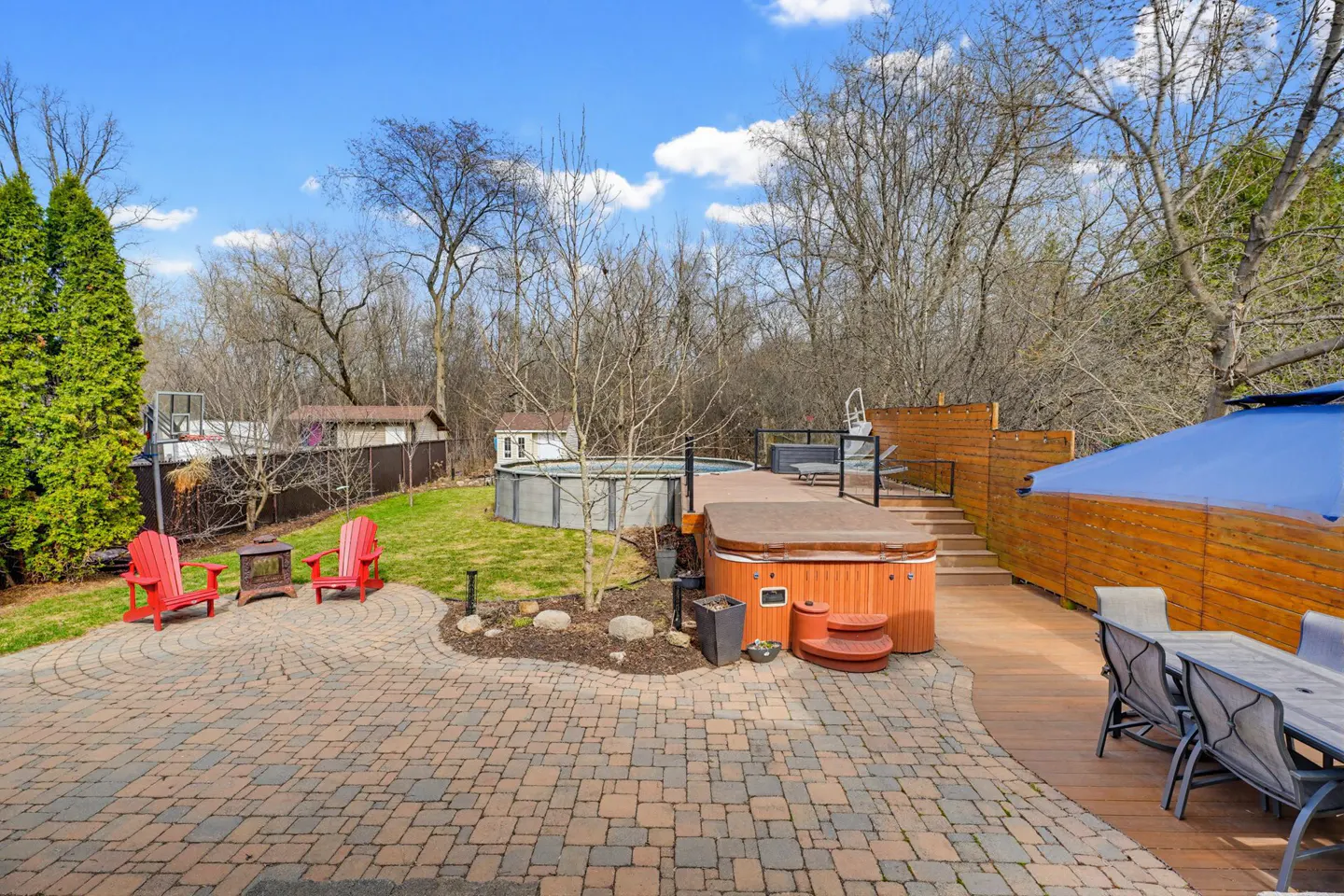 Backyard patio with red chairs, a hot tub on a wooden deck, an above-ground pool, and a dining set under a blue umbrella.