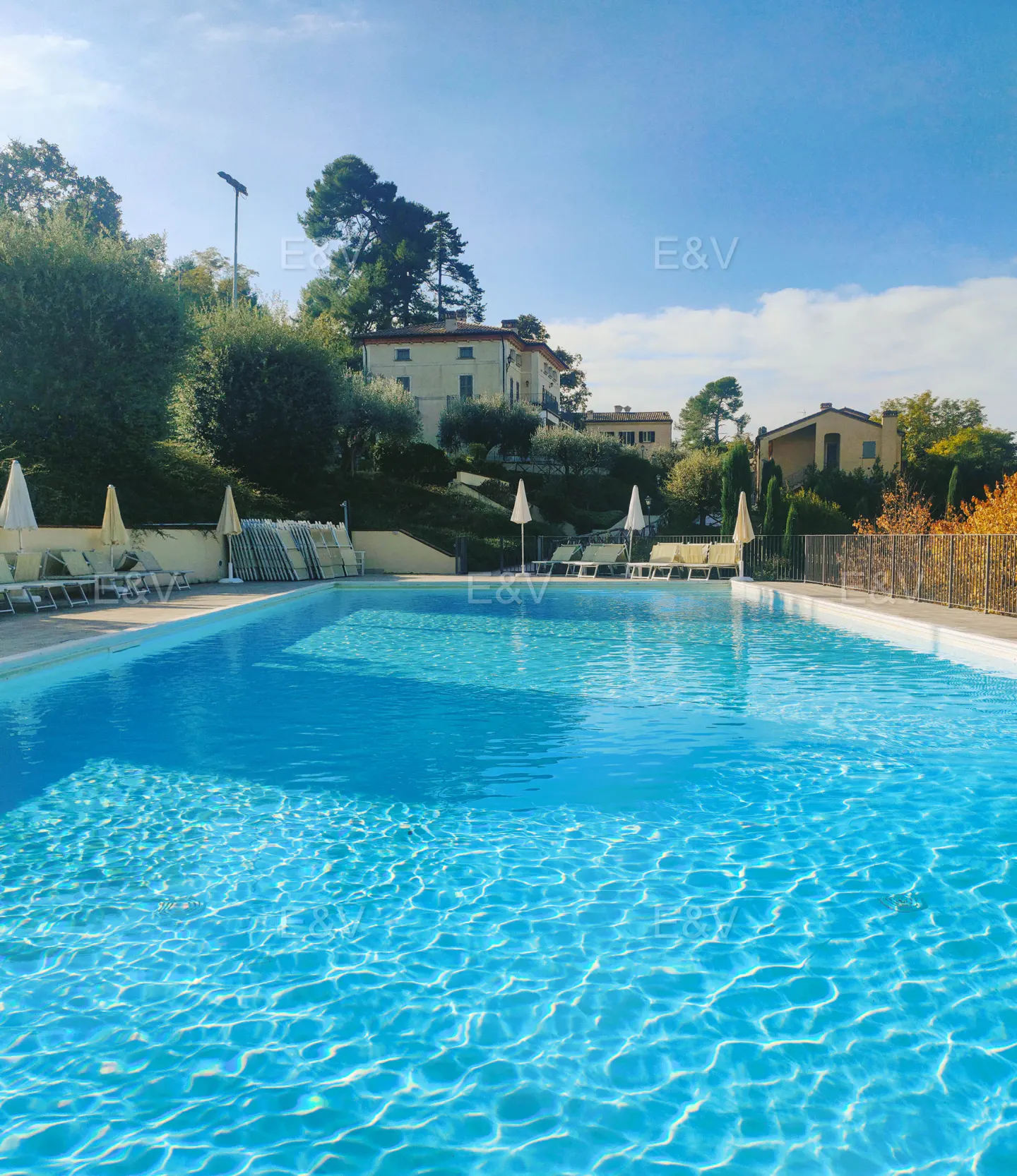 A bright blue swimming pool with lounge chairs and umbrellas, set against a backdrop of trees and buildings.