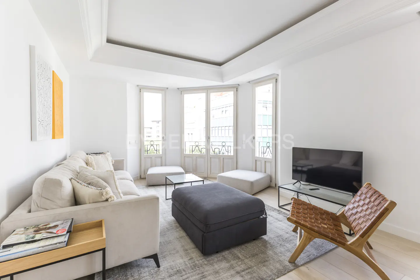 Bright living room with white walls, light wood floors, and a gray rug. A beige sofa, ottoman, and woven chair furnish the space. Three windows offer a city view.