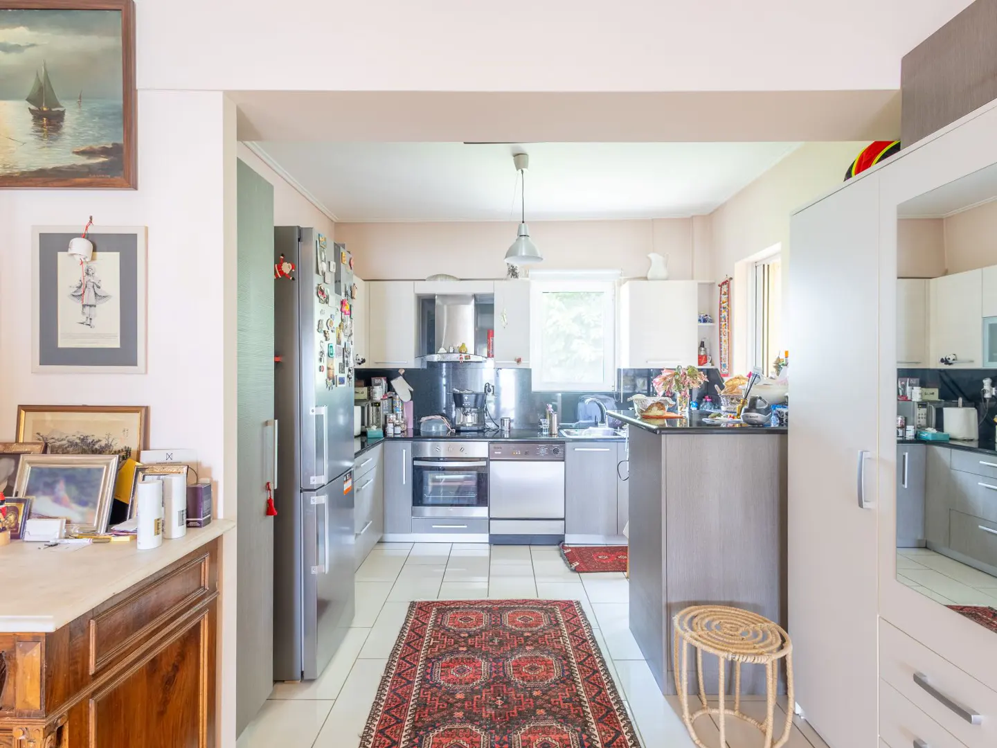 A bright kitchen with white cabinets, stainless steel appliances, and a red patterned rug on white tile floor.