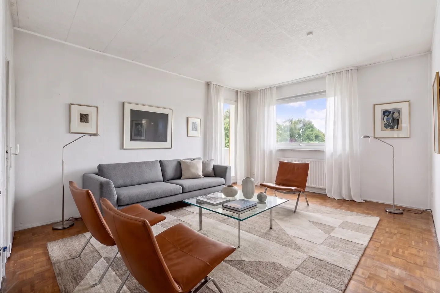 Bright living room with a gray sofa, two brown leather chairs, and a glass coffee table on a geometric rug. White walls and hardwood floors.
