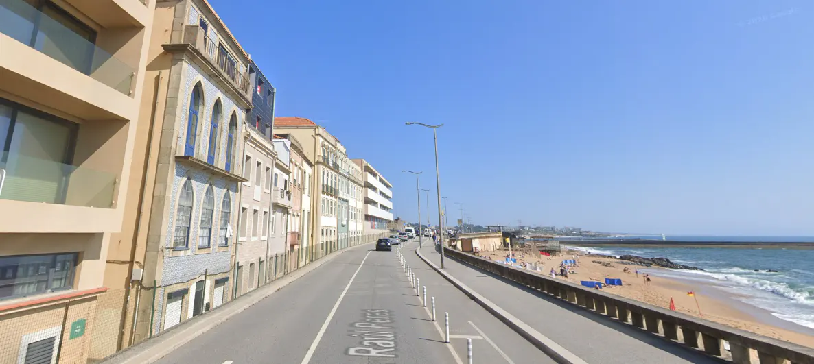 Coastal road view with buildings on the left and a beach on the right under a clear blue sky.