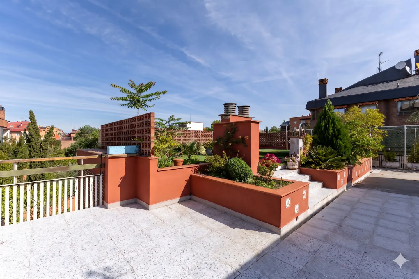 A rooftop terrace with terracotta planters, greenery, and a white tiled floor under a blue sky.