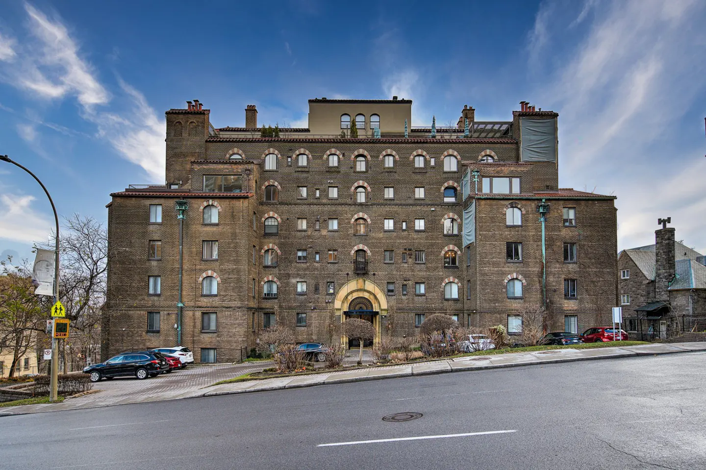 Exterior view of a large, brown brick apartment building with arched windows and a central arched entrance. Cars are parked in front.