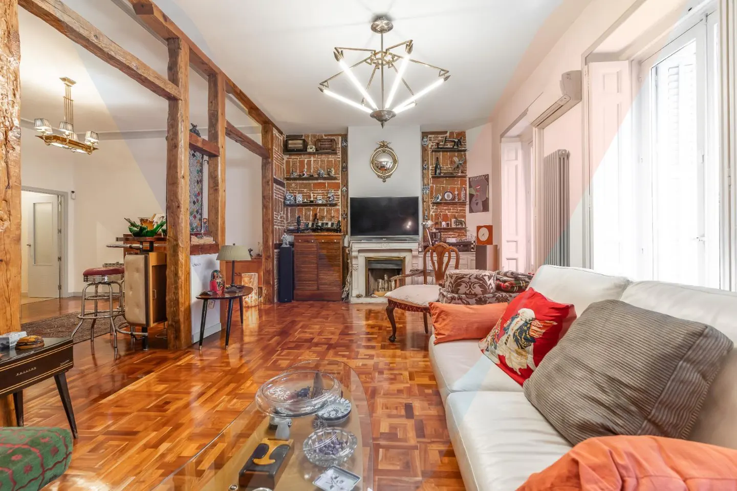 Living room with wood floors, white sofa, and exposed brick. A modern chandelier hangs above a TV over a fireplace.