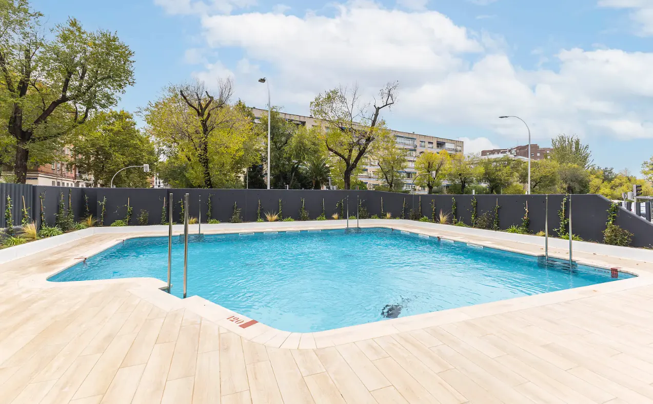Outdoor swimming pool with clear blue water, light wood-look decking, and a dark privacy fence against a backdrop of trees.