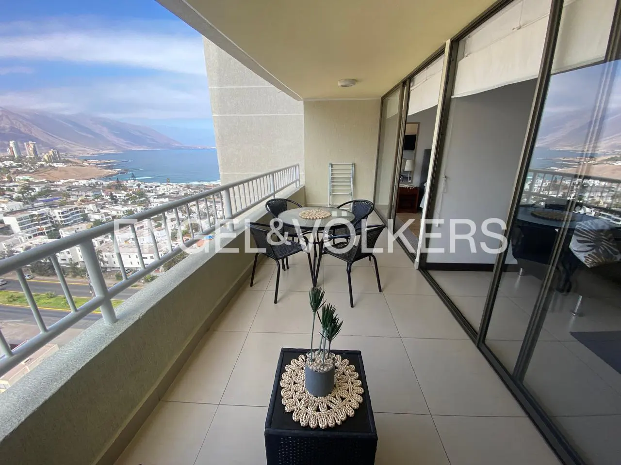 Balcony with a table and chairs overlooking a coastal city and ocean. Sliding glass doors lead to the interior.