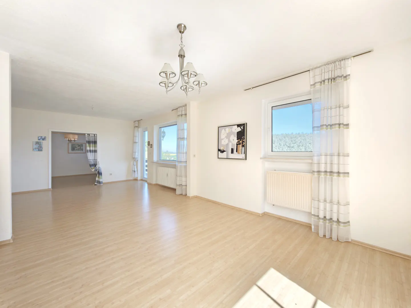 Bright, empty living room with light wood floors, white walls, and a chandelier. Windows with sheer curtains let in natural light.