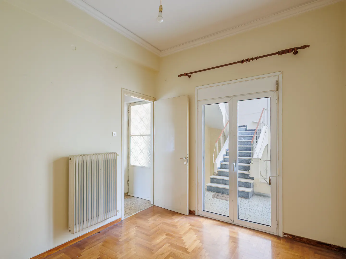Empty room with light walls, herringbone wood floors, a radiator, and glass doors looking out to an indoor staircase.