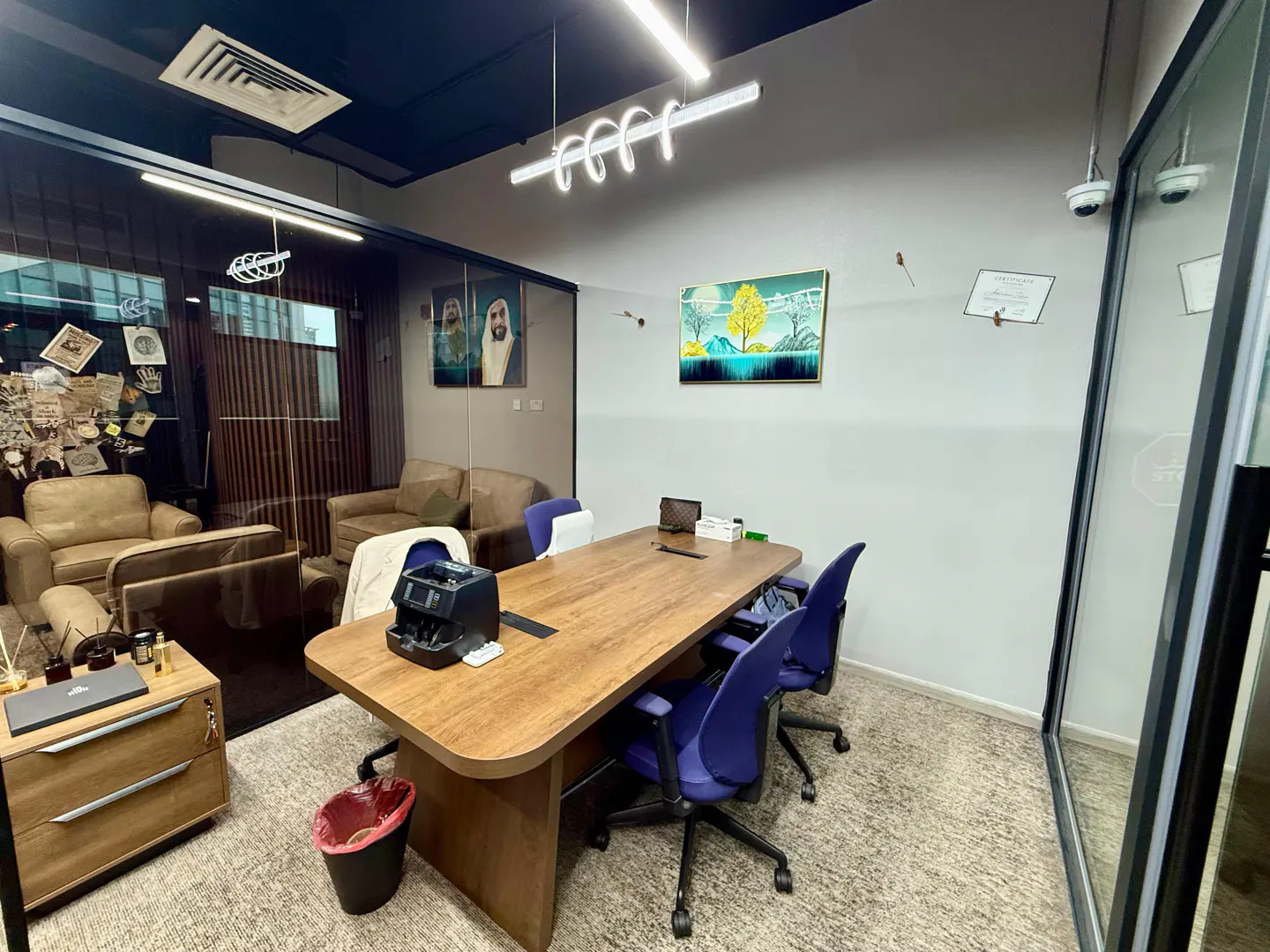 Office interior with a long wooden table, blue chairs, and tan sofas. Artwork and portraits adorn the walls. A modern light fixture hangs above.