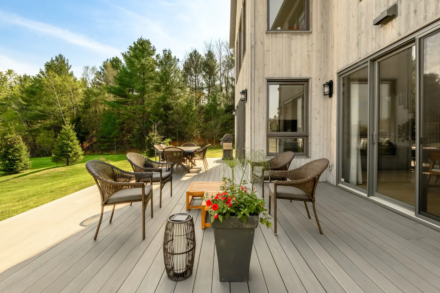 Outdoor patio with wicker furniture, a wooden table, and a potted plant on a gray deck. A modern house and green trees are in the background.