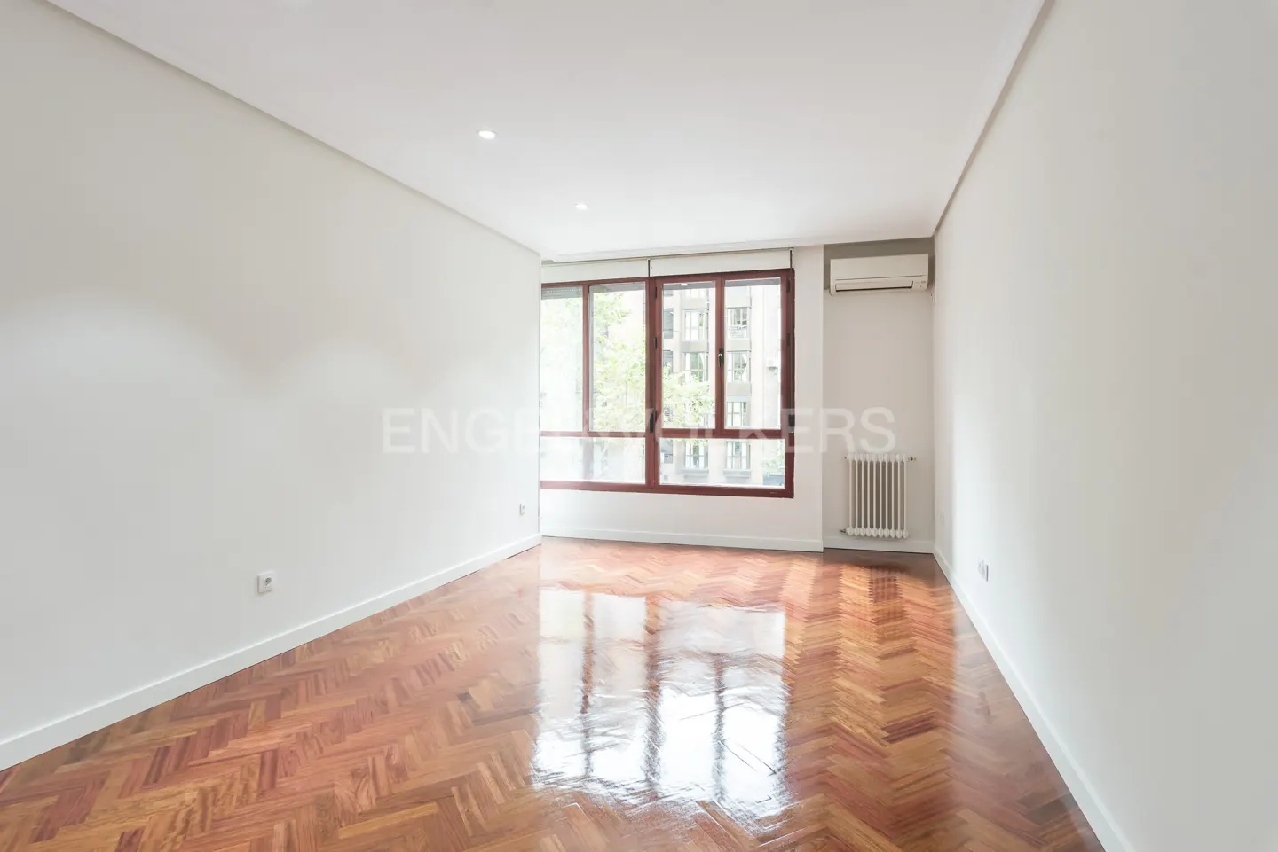 Empty room with herringbone wood floor, white walls, large window with brown frame, radiator, and air conditioner.