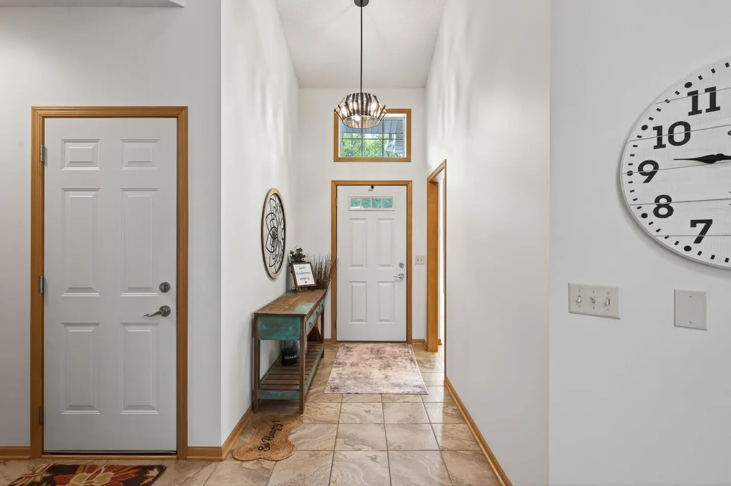 Entryway with white walls, tile floor, and a console table. A white door is visible at the end of the hall. A clock hangs on the right wall.