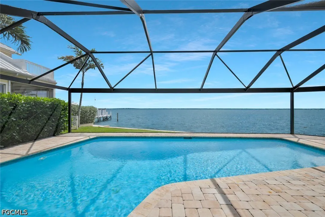 Screened pool with blue water and brick patio overlooks a bay under a blue sky. A dock is visible in the distance.