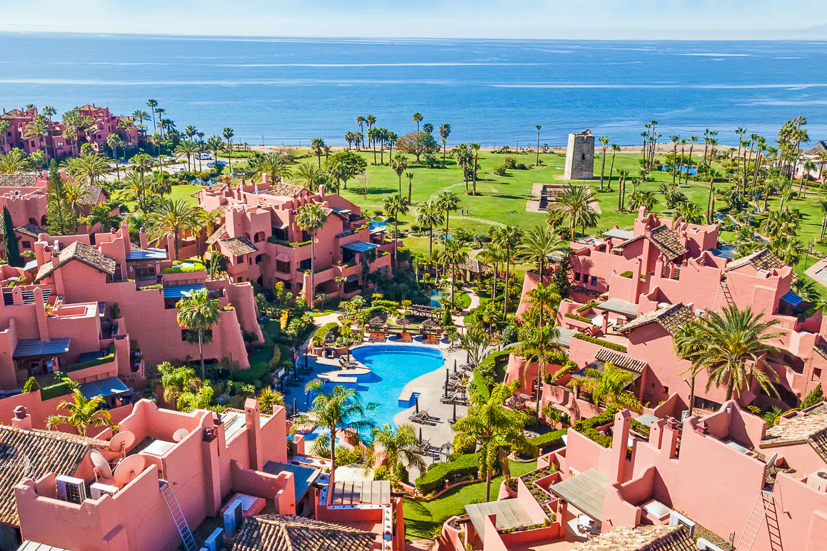 Aerial view of a pink resort complex with a pool, palm trees, and the ocean in the background.