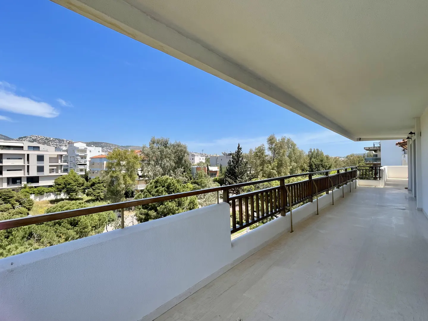 Wide balcony with white walls and brown railings overlooking trees, buildings, and a blue sky.