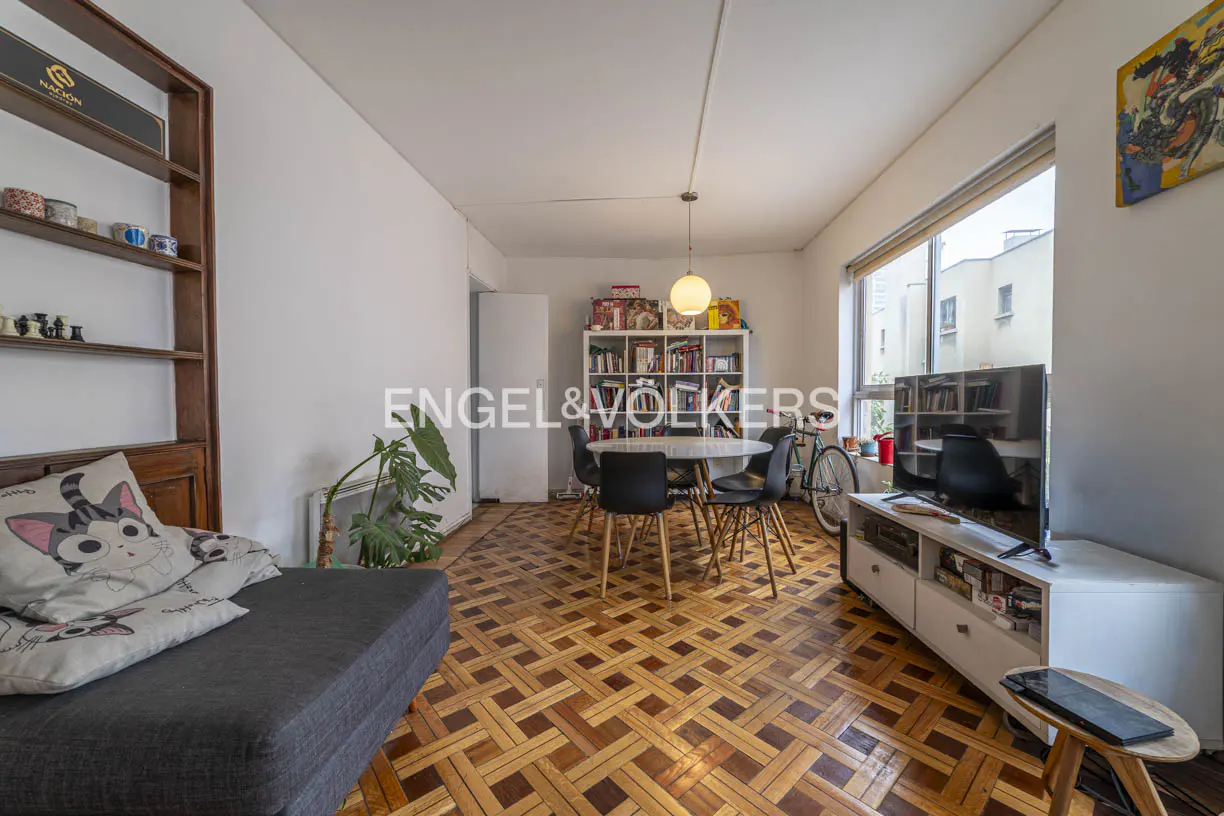 Living room with parquet floors, a grey sofa, dining table with black chairs, TV on a white stand, and a bookshelf.