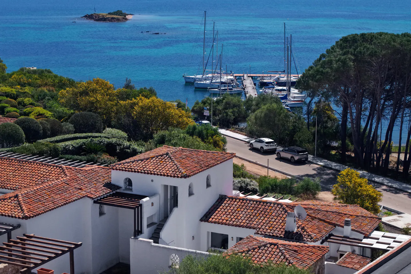 View of white villas with red tile roofs overlooking a marina with sailboats and turquoise water.