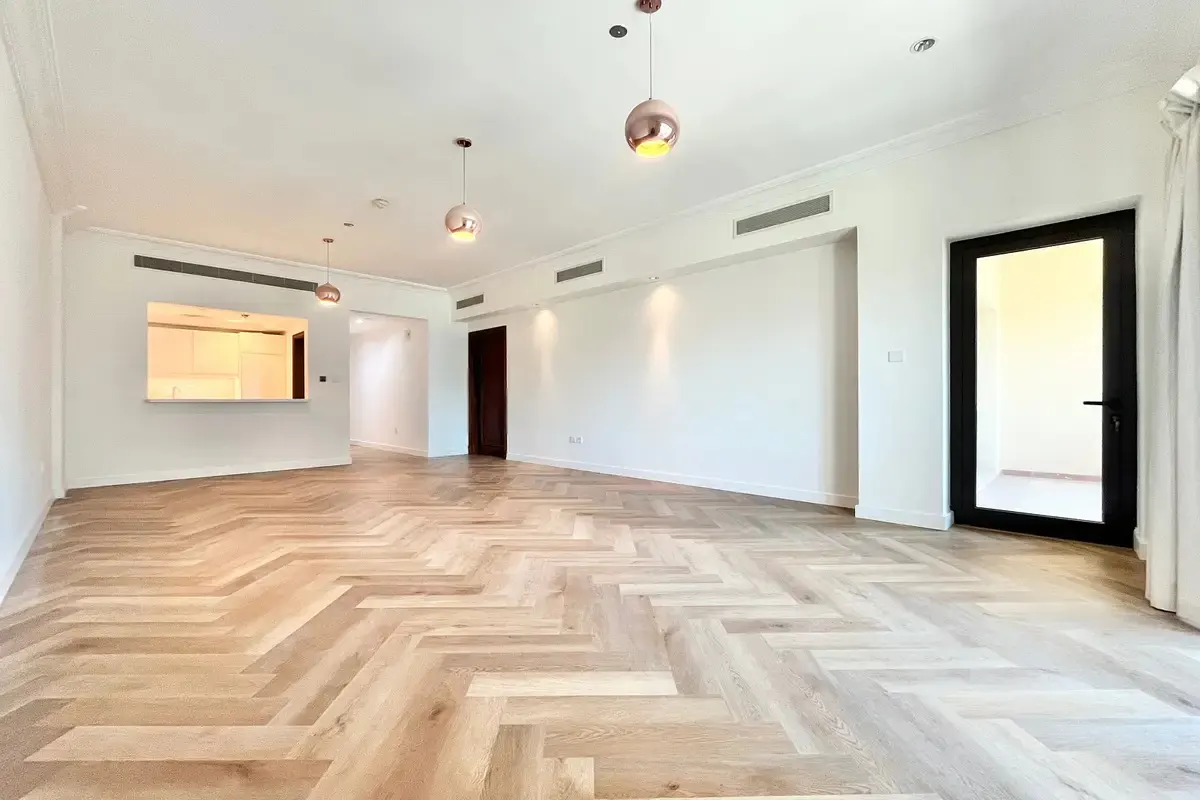 Bright, empty room with herringbone wood floors, white walls, and three rose gold pendant lights. A black door and kitchen pass-through are visible.