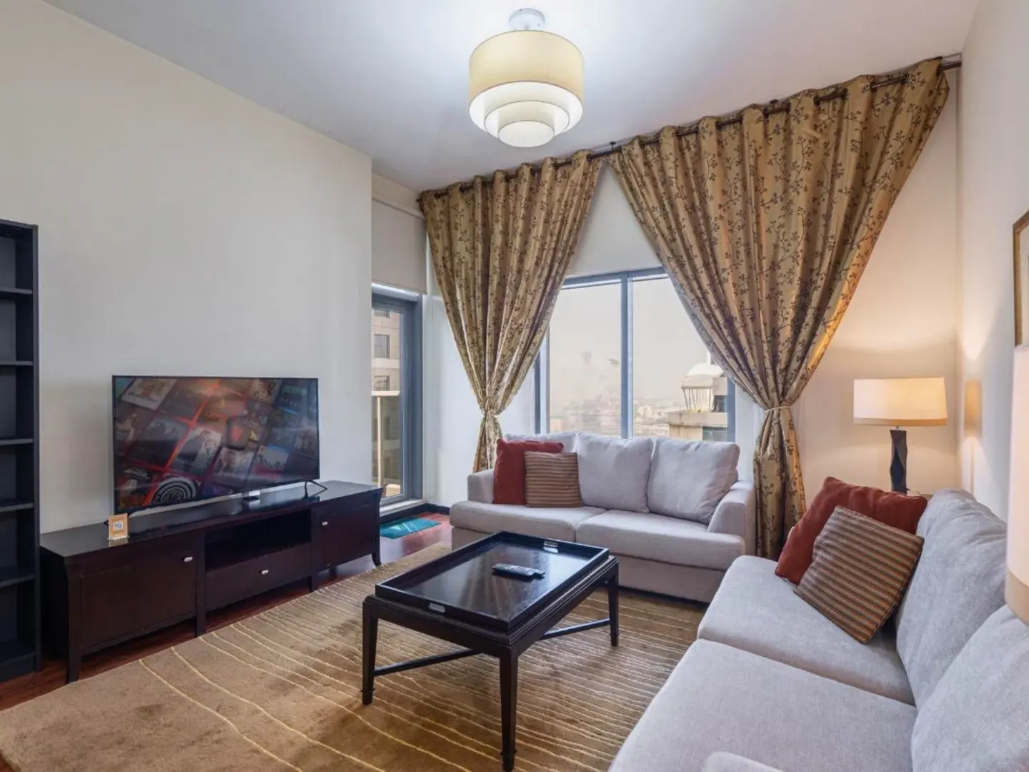 Living room with beige sofas, brown patterned curtains, and a dark wood coffee table on a striped rug. A TV sits on a dark stand.