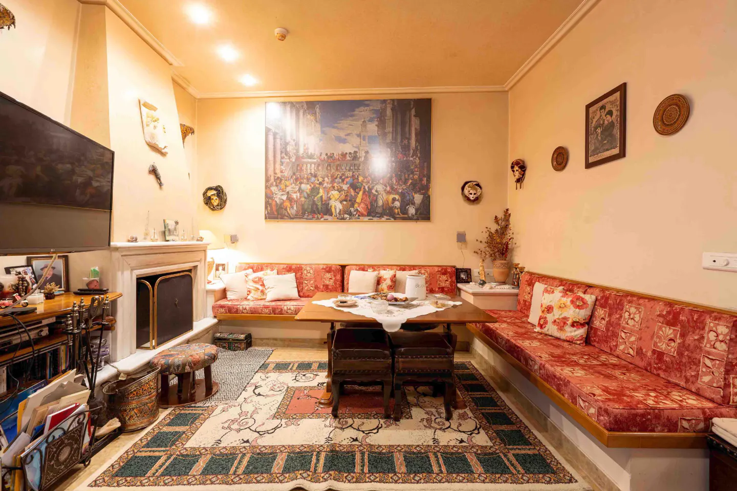 Living room with built-in red floral couches, wooden table, and fireplace with TV. A large painting hangs above the couch.