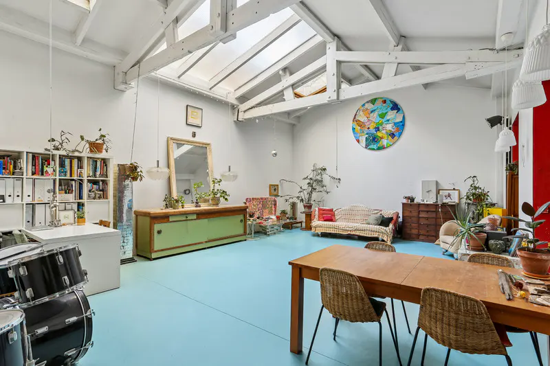 Bright, airy loft apartment with blue floors, white walls, and skylights. A dining table, sofa, and drum set furnish the space.