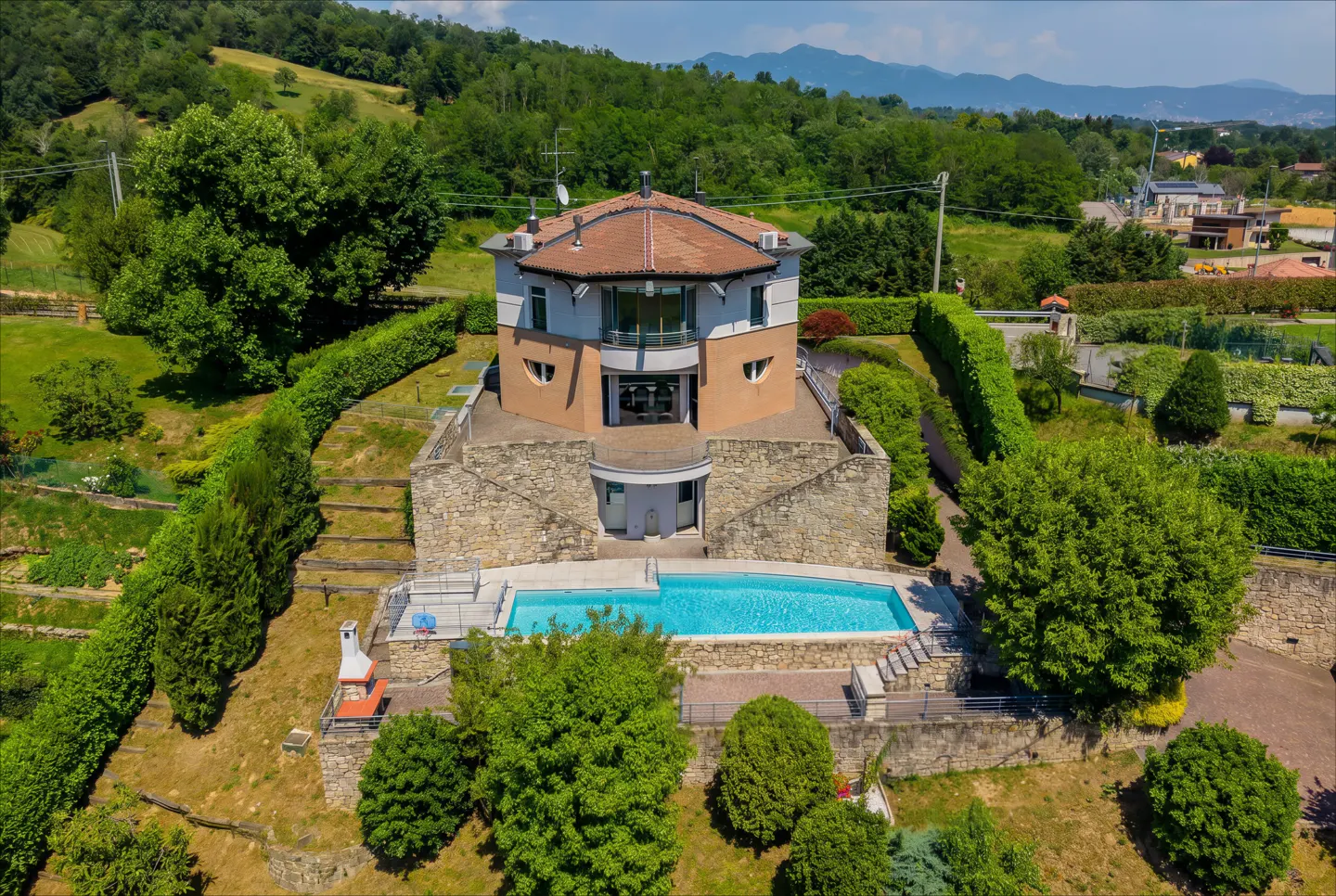 Aerial view of a multi-story home with a stone base, tan upper levels, and a swimming pool set in a lush, hilly landscape.