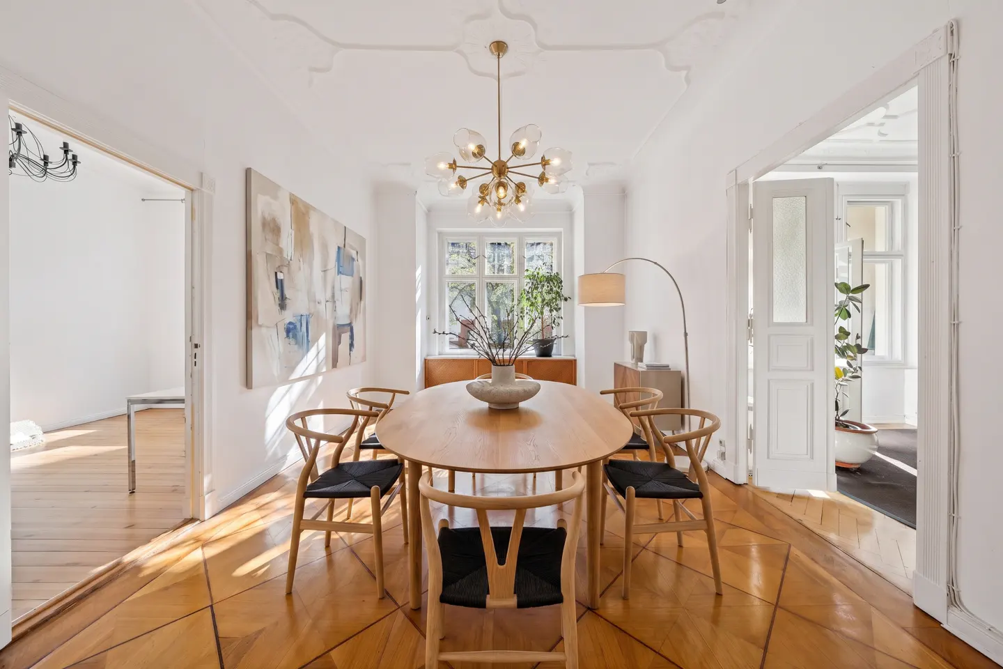 Bright dining room with wood floors, white walls, and an oval table surrounded by six chairs. A modern chandelier hangs above the table. Artwork and plants add to the room's decor.