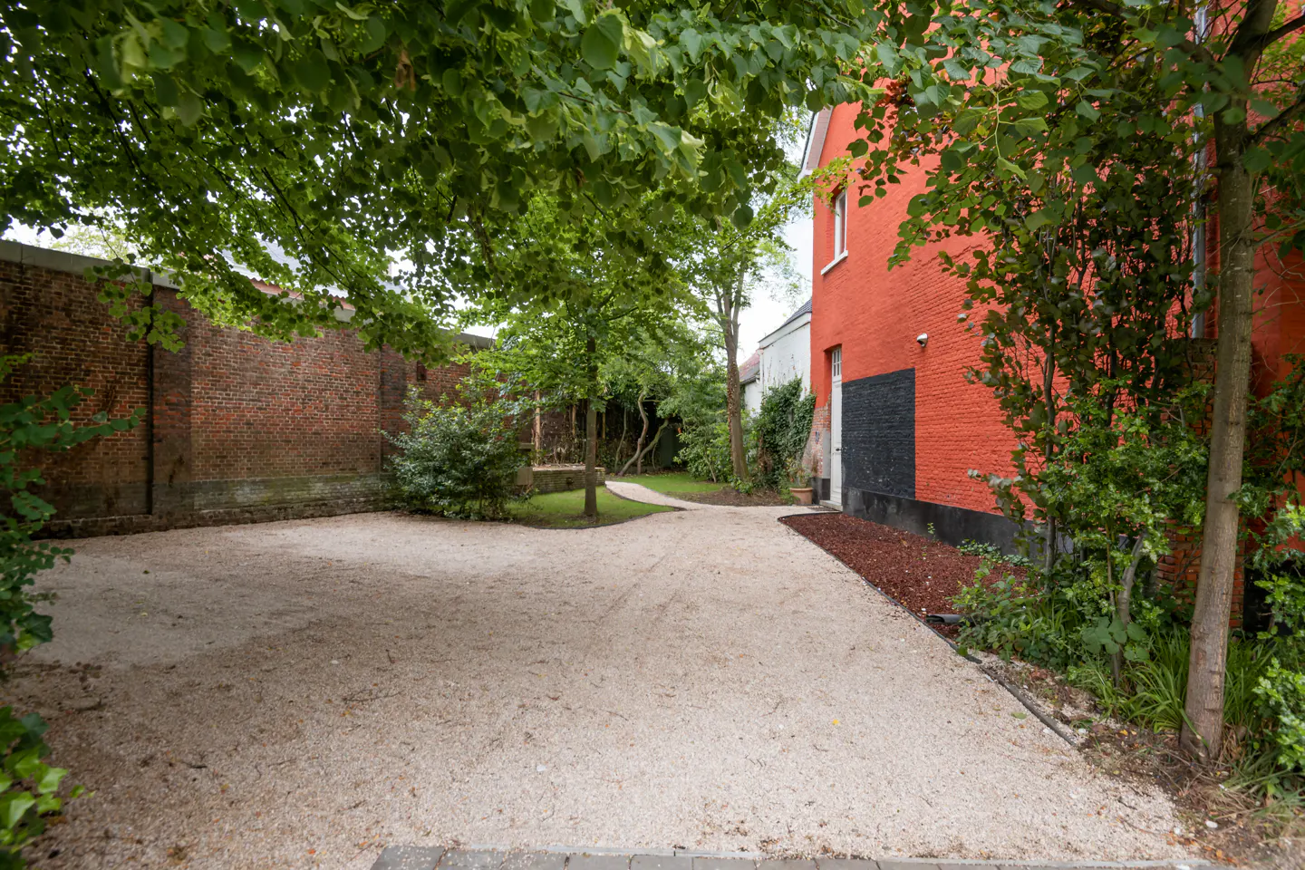 A gravel driveway leads past a bright red brick building toward a garden path, framed by lush green trees and brick walls.
