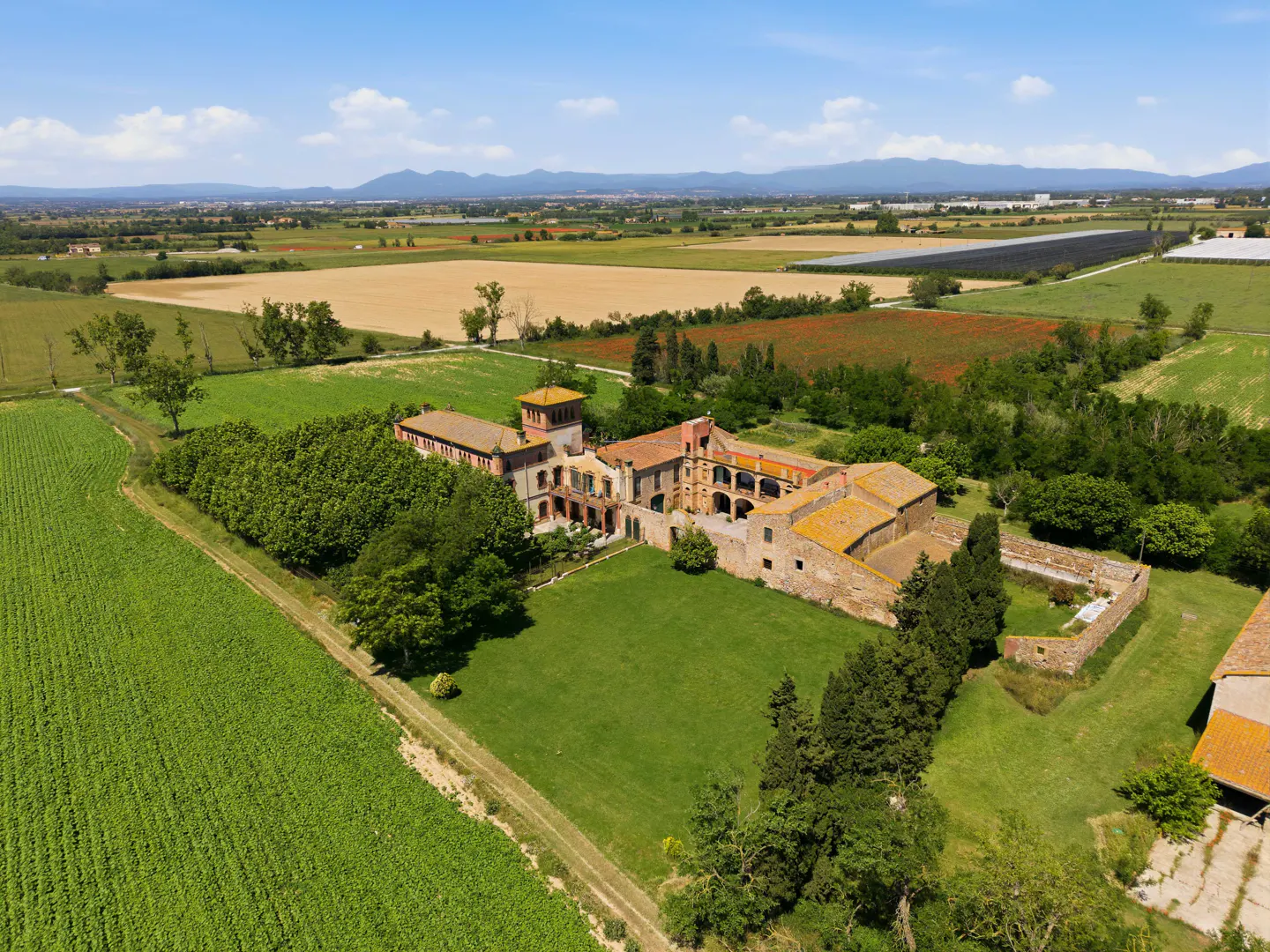 Aerial view of a large, red-roofed stone farmhouse surrounded by green fields and trees under a blue sky.