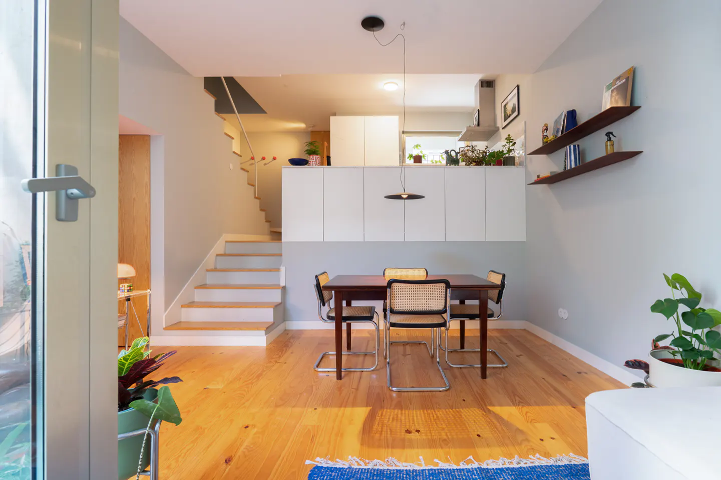 Bright, modern dining room with wood floors, a dark wood table, and four chairs. White cabinets and shelves line the wall. Stairs lead to an upper level.