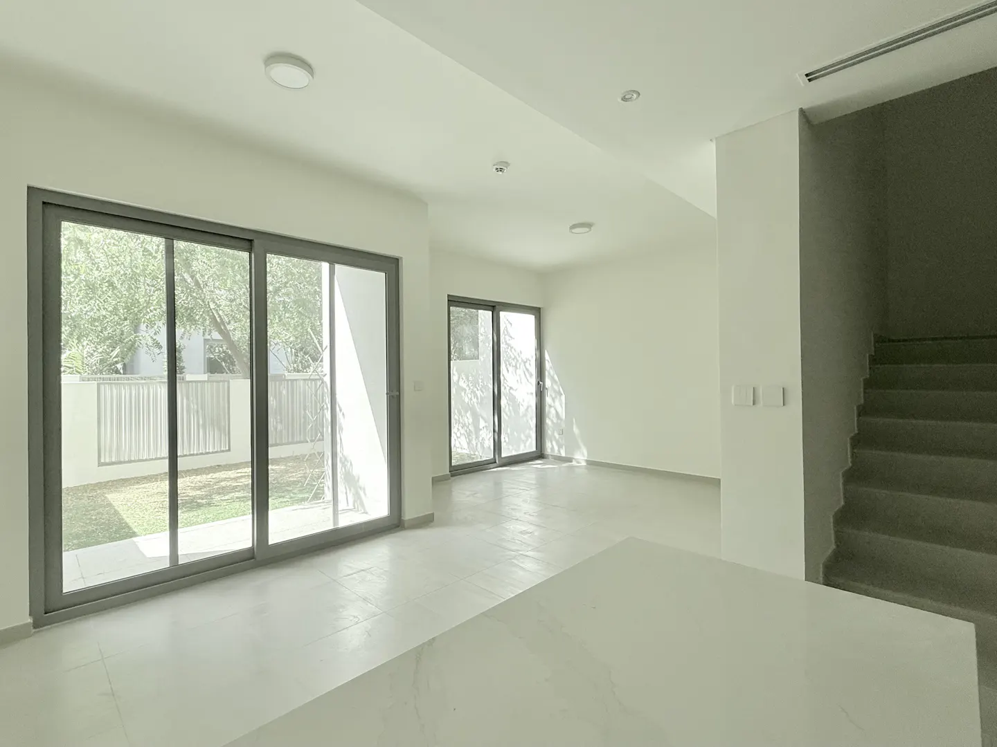 Bright, empty room with white walls, tile floor, and stairs. Sliding glass doors lead to a yard with a fence and trees.
