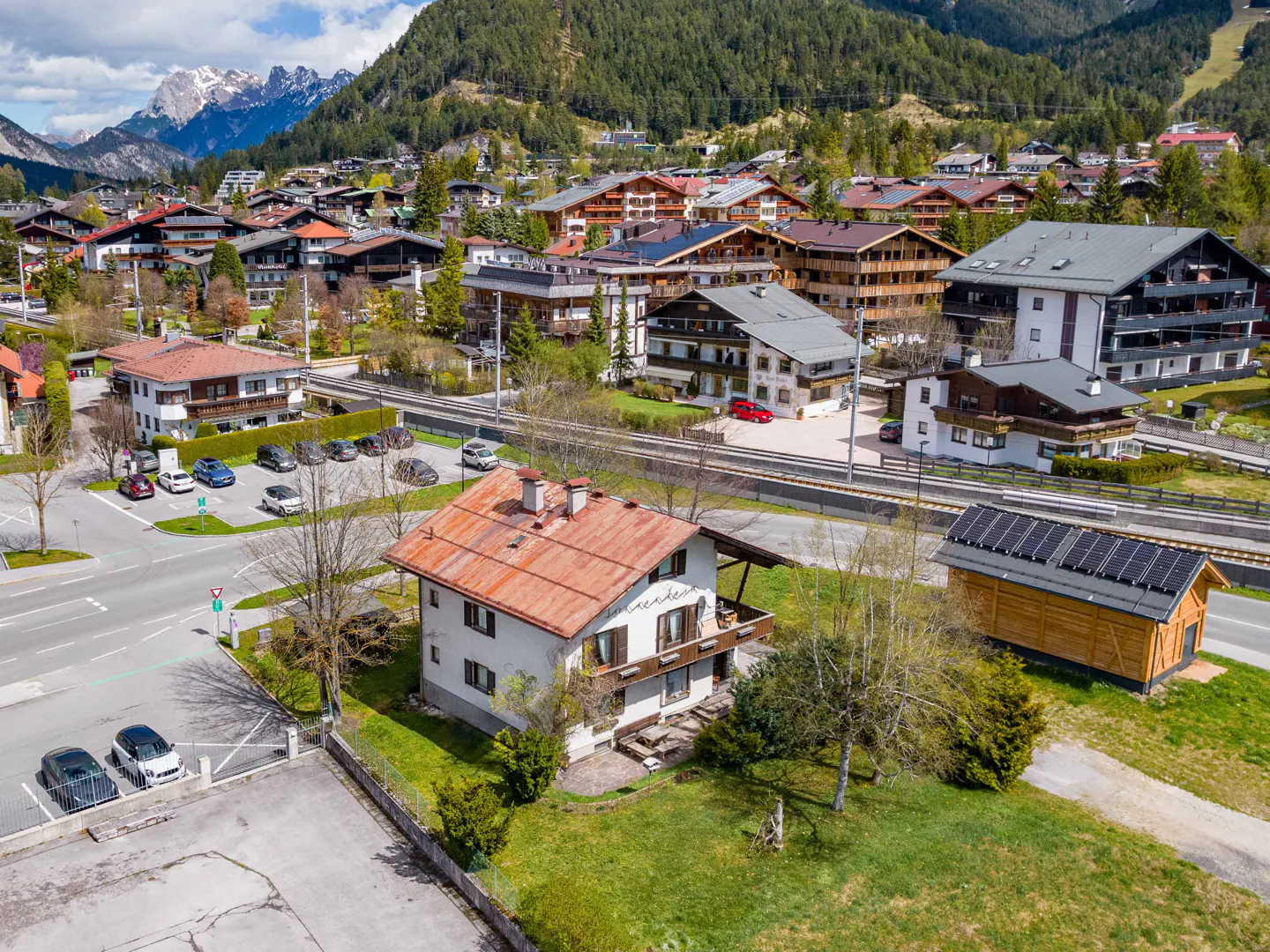 Aerial view of a European town with houses, trees, and mountains in the background. A train track runs through the town.