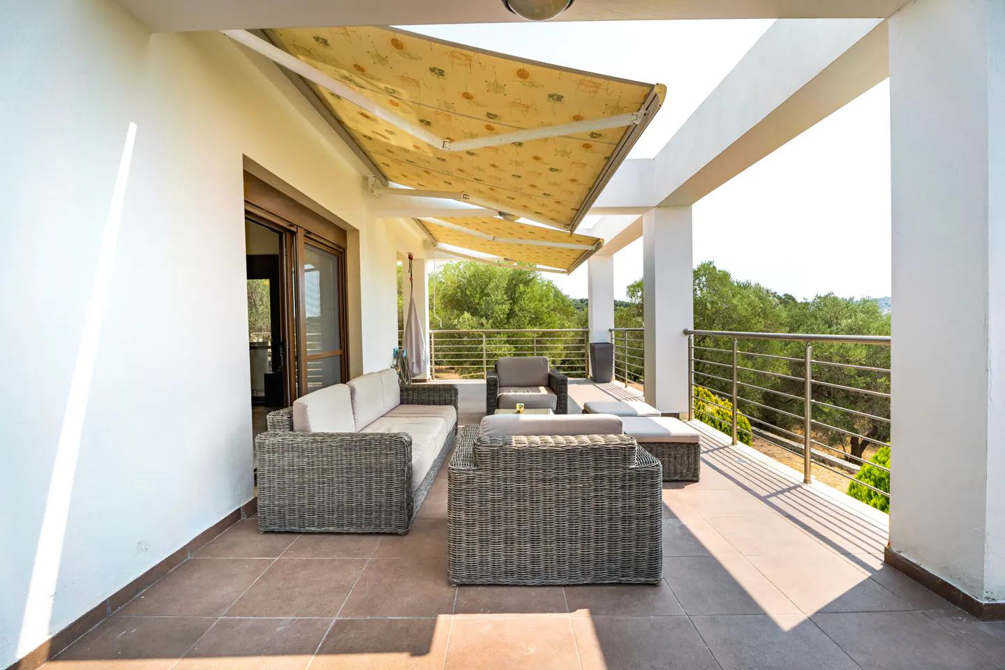 Outdoor patio with wicker furniture, including a sofa, chairs, and ottomans, under a yellow awning. A metal railing overlooks green trees.