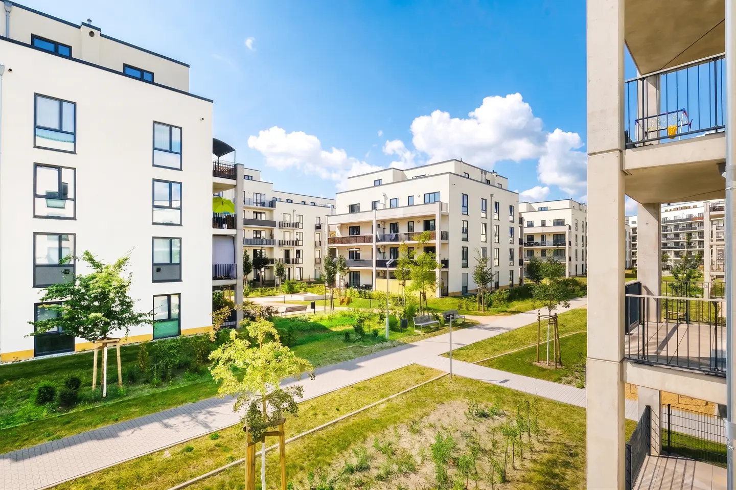 Modern white apartment buildings with balconies surround a green courtyard under a blue sky.