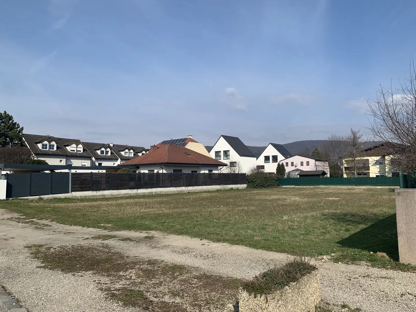 Vacant lot with patchy grass in a residential area. Houses with varied rooflines and colors line the background under a blue sky.