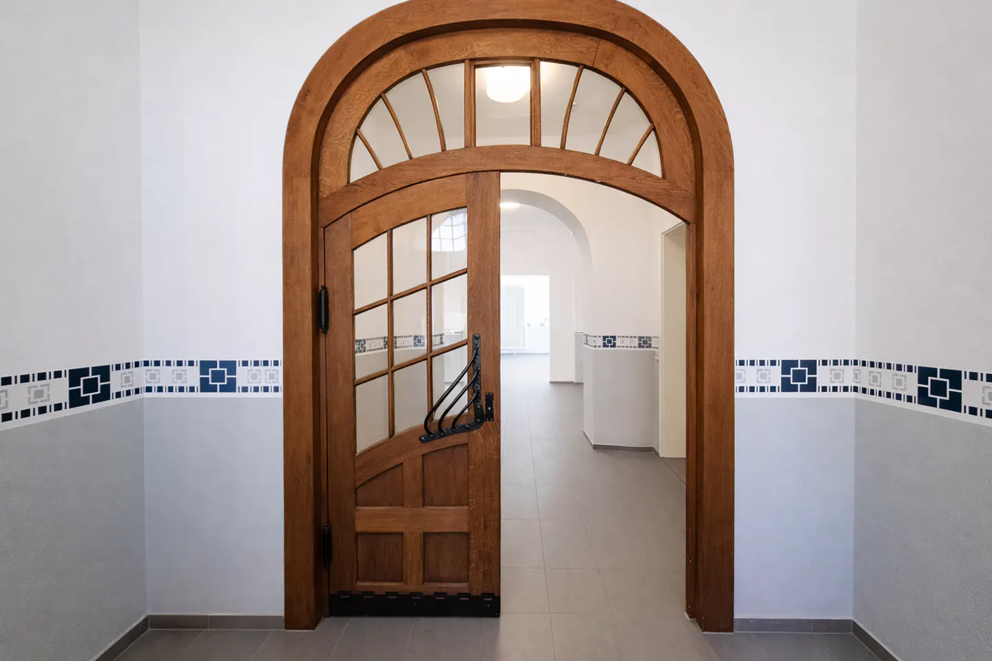 Arched wooden door with glass panes, slightly ajar, in a white hallway with blue patterned trim.