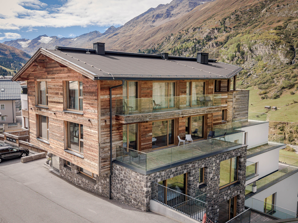 Modern wooden chalet with stone base, glass balconies, and mountain views in a scenic alpine setting. [AI-generated text]