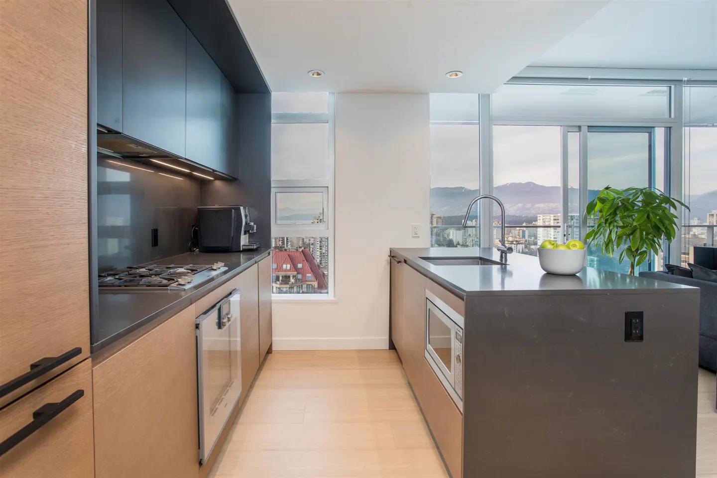 A modern kitchen with wood cabinets, dark countertops, and stainless steel appliances. A large window offers a city view.