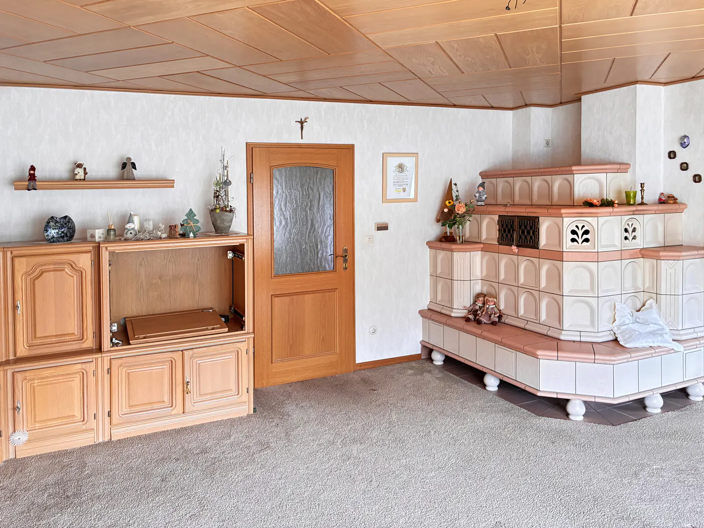 Living room with wood paneled ceiling, light carpet, wooden cabinets, door, and a large tiled stove with pink trim.